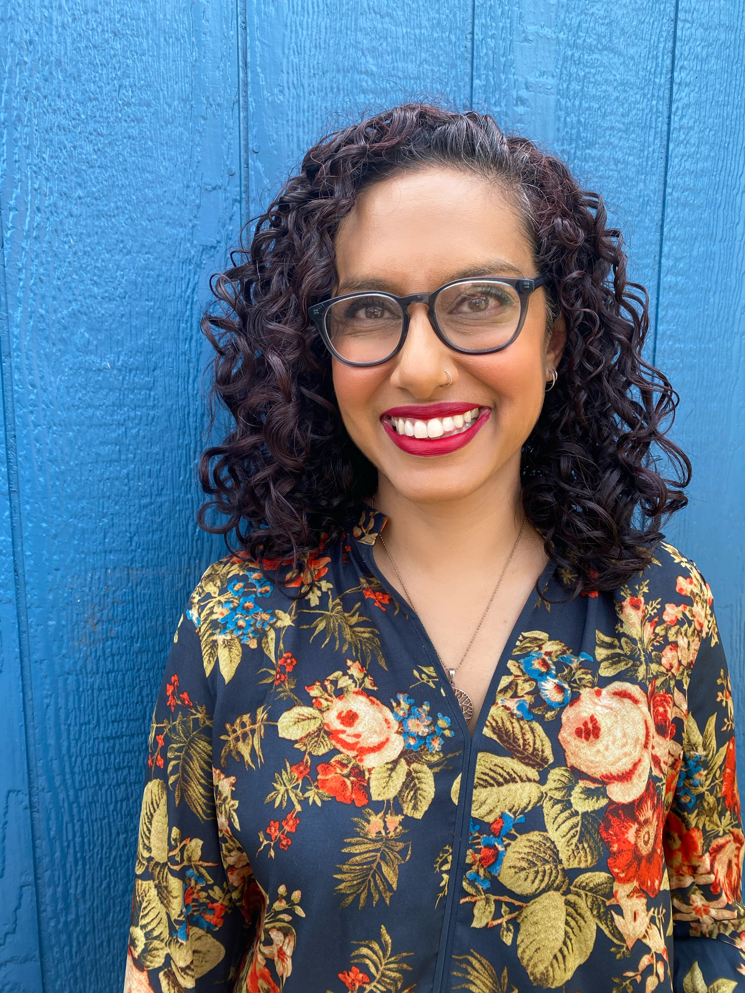 Picture of Farah-- A south Asian woman with black curly hair reaching her shoulders and brown skin. She is wearing a blue shirt with large roses on it, and standing in front of a blue wall. 
